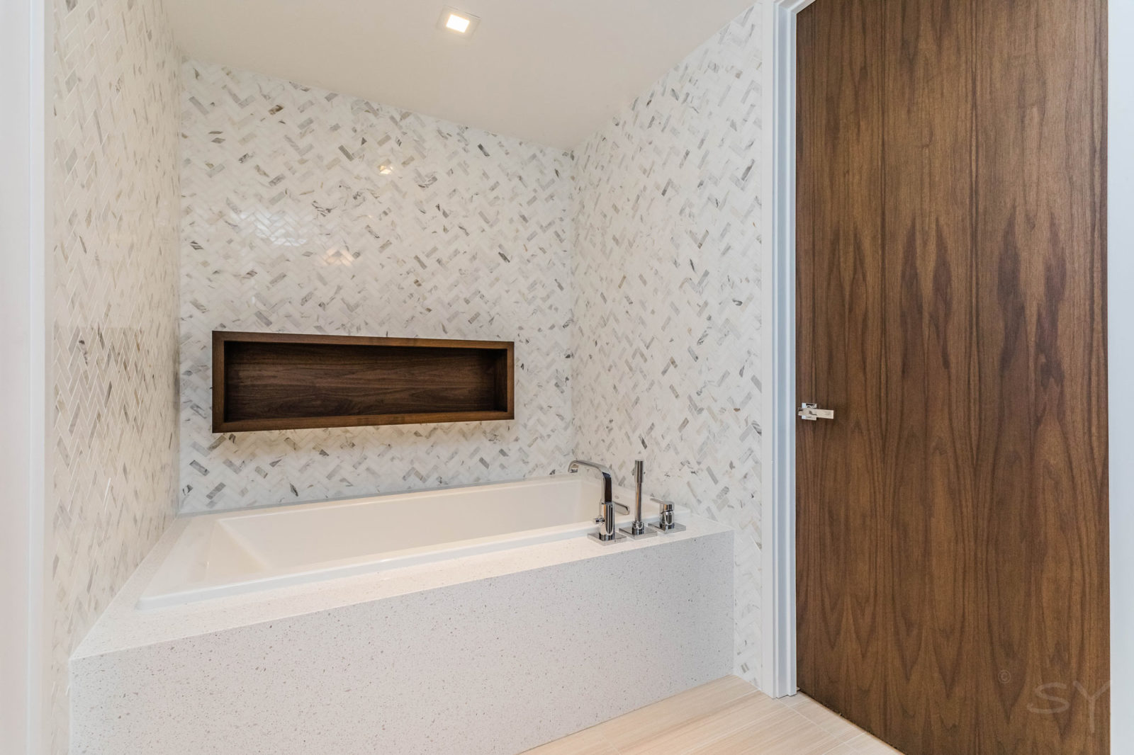 large built in bathtub with light patterned tile walls dark wooden door and chrome finishes