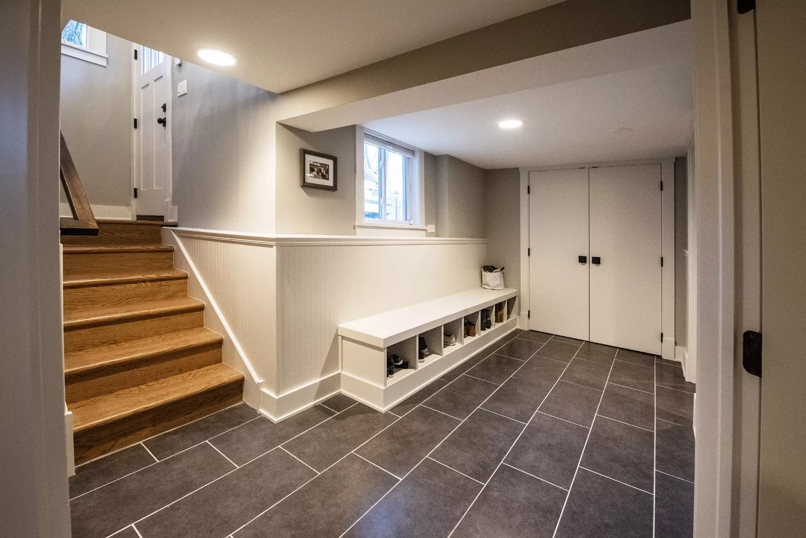Hallway leading from exterior doors with grey tiled floor & bench with shoes on shelves underneath