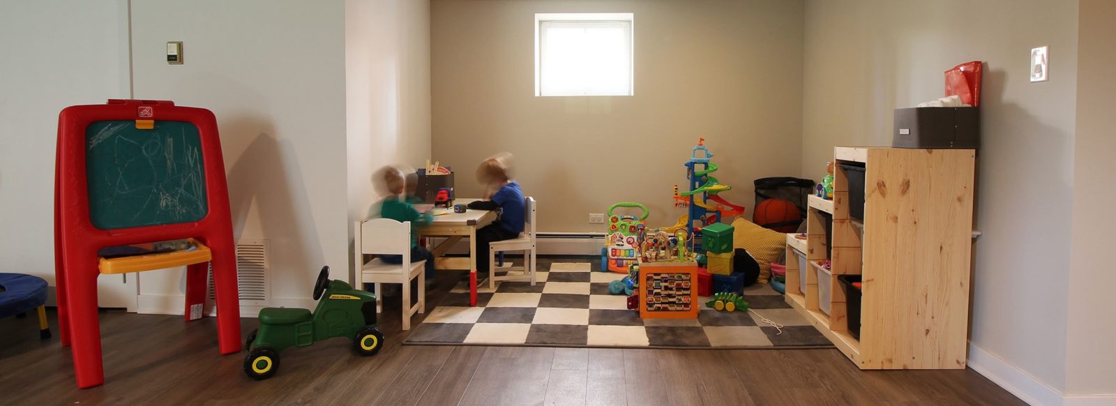 two children playing at a table in a basement playroom surrounded by other toys on a patterned rug