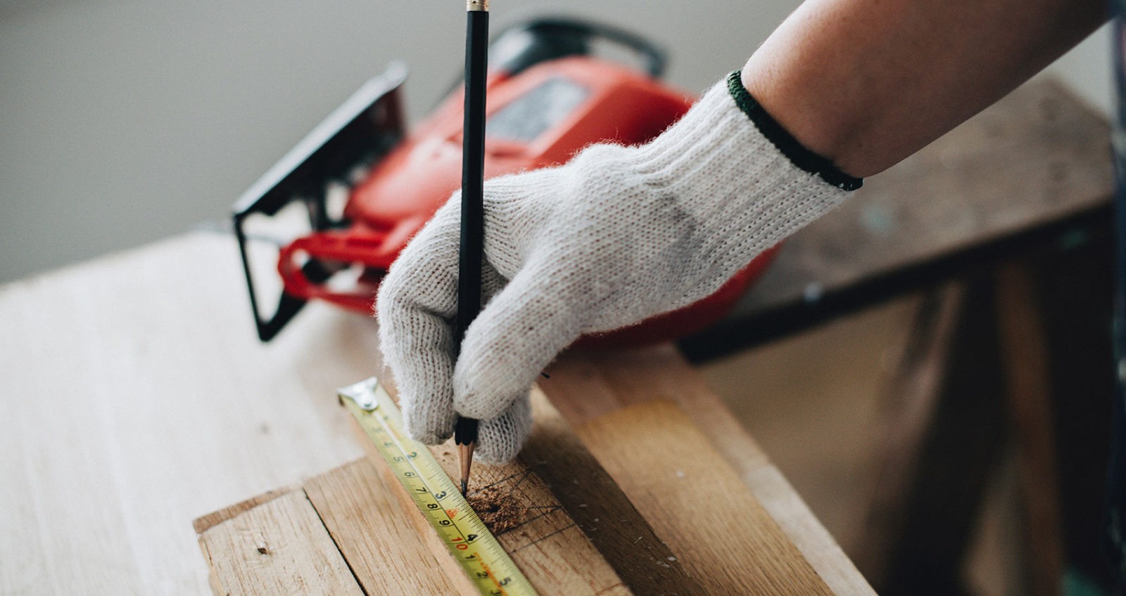 hand with glove holding pencil against piece of wood and tape measurer next to a power saw