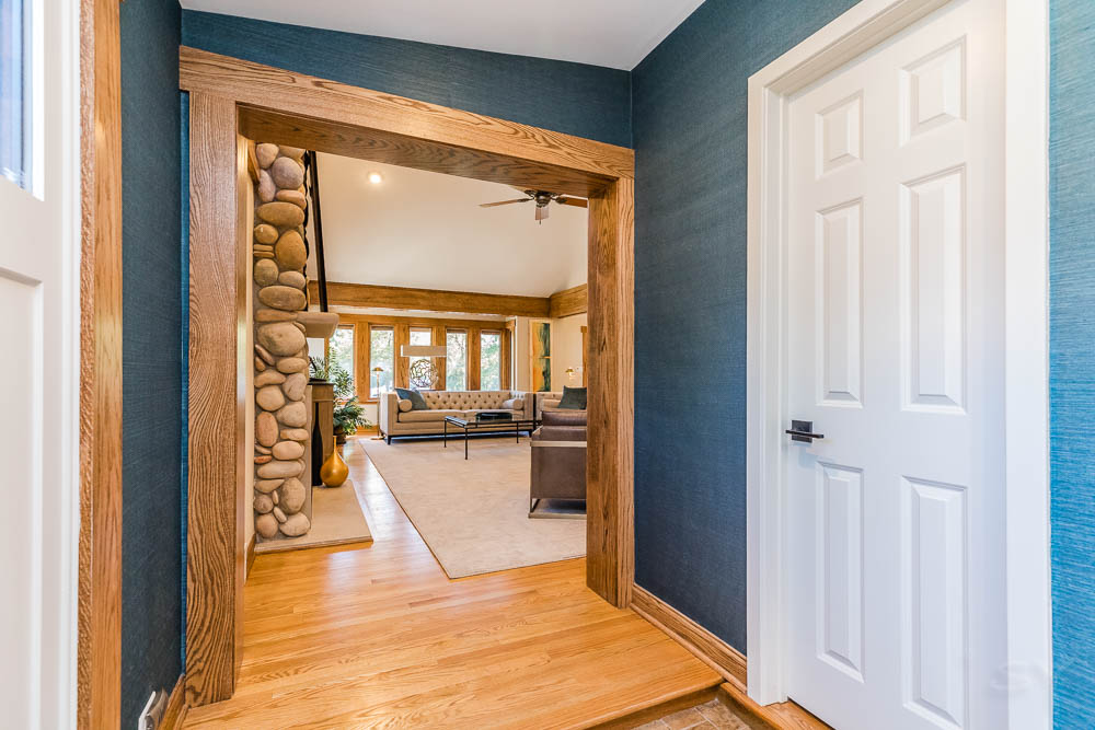 Hallway looking into a living room with tan cobblestone fireplace, couch & chairs, windows