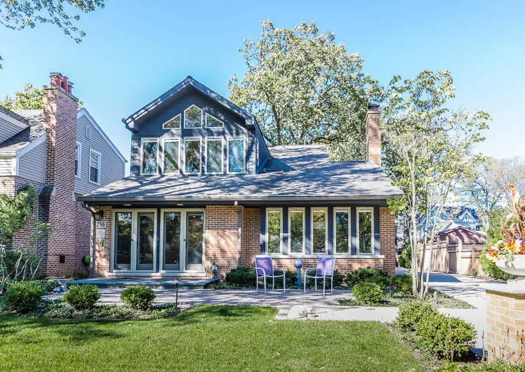 two story brick and blue home with many windows two chairs on back patio and green yard with shrubs