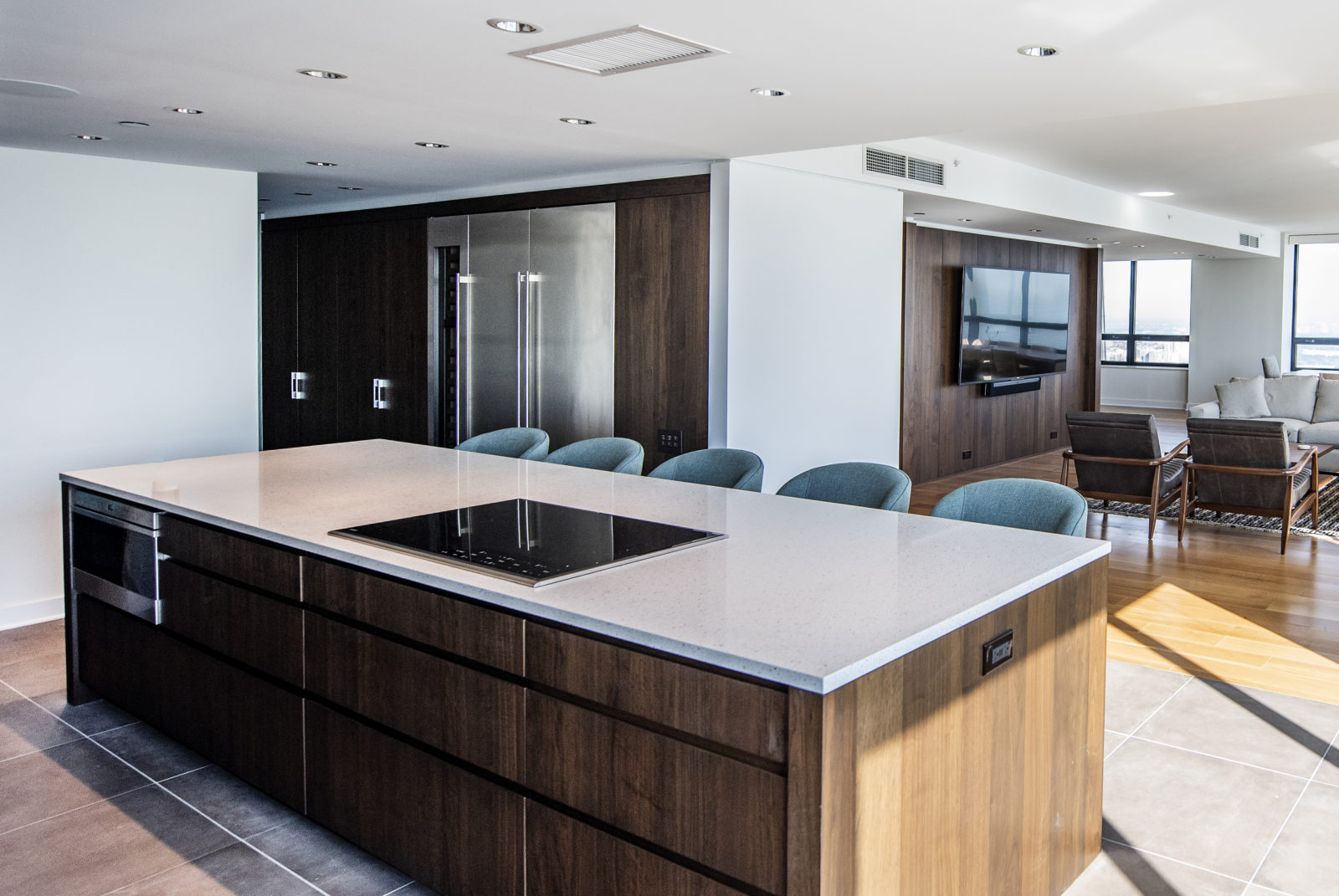 Kitchen with island & chairs, built-in oven & flat stovetop, adjacent to living room with couch & TV