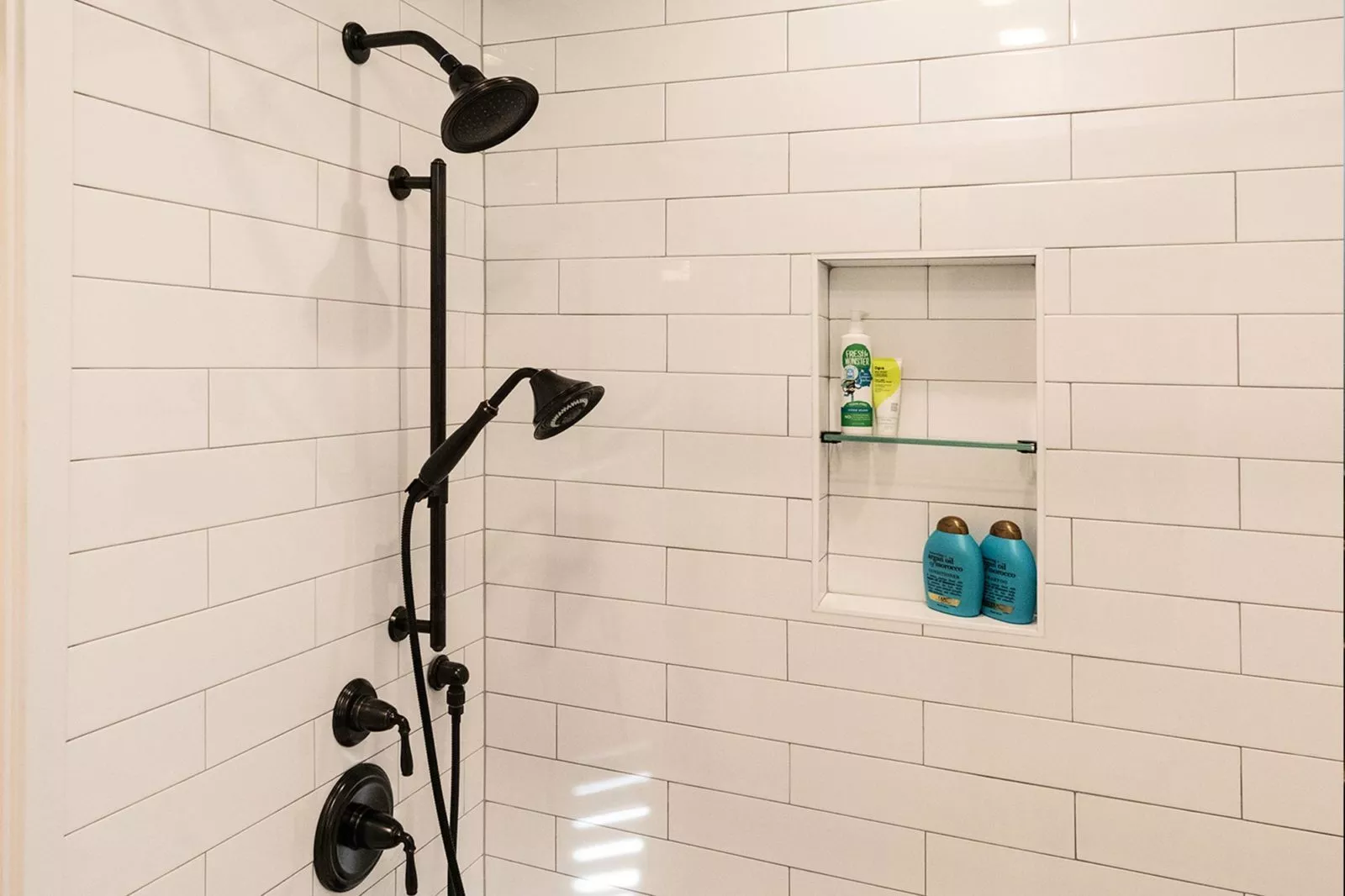 Inside of a shower with white tile walls, black shower head & handles, and products on shelf