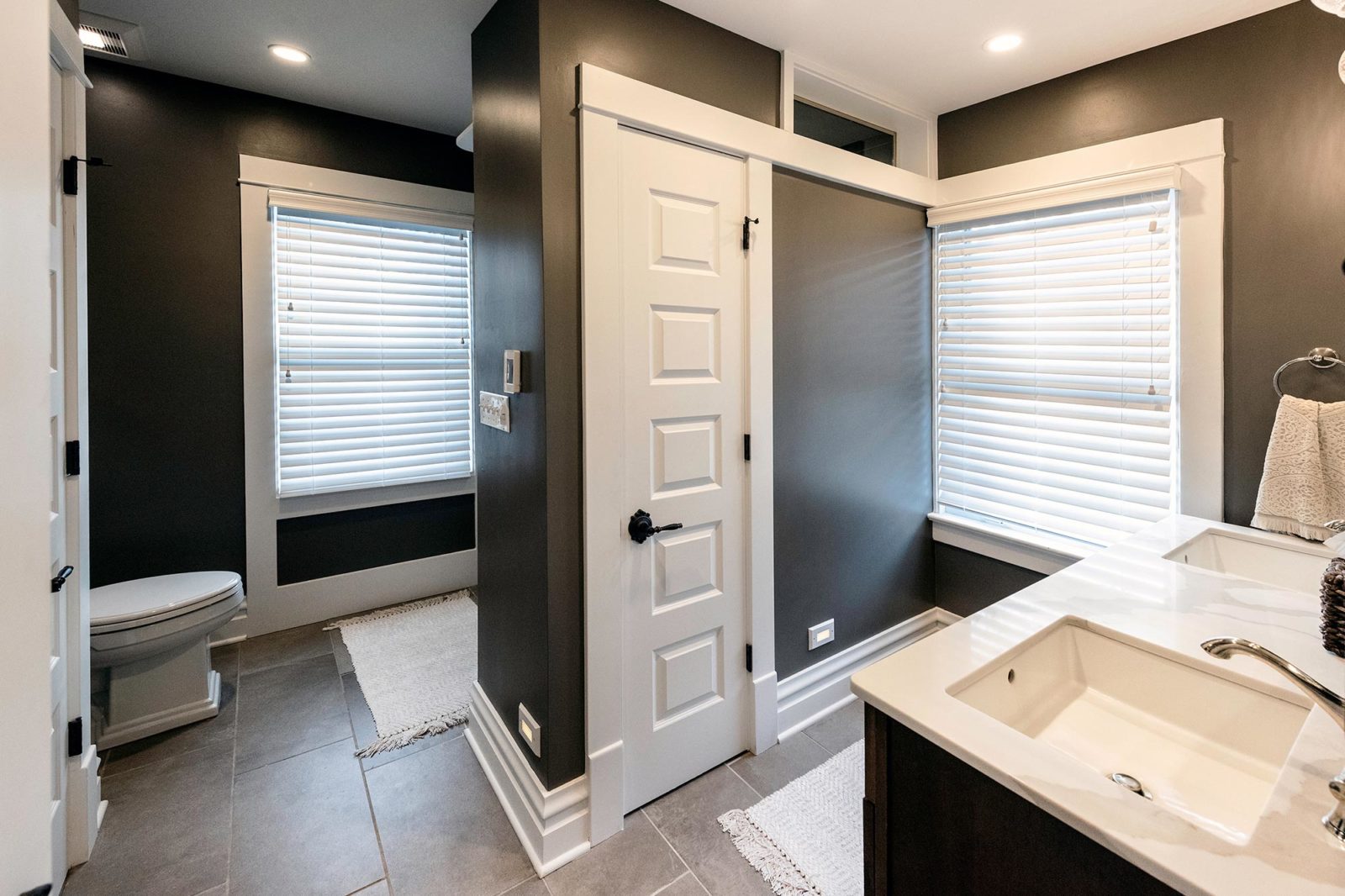 U-shaped bathroom with grey walls, toilet, white sinks, small closet, two exterior windows