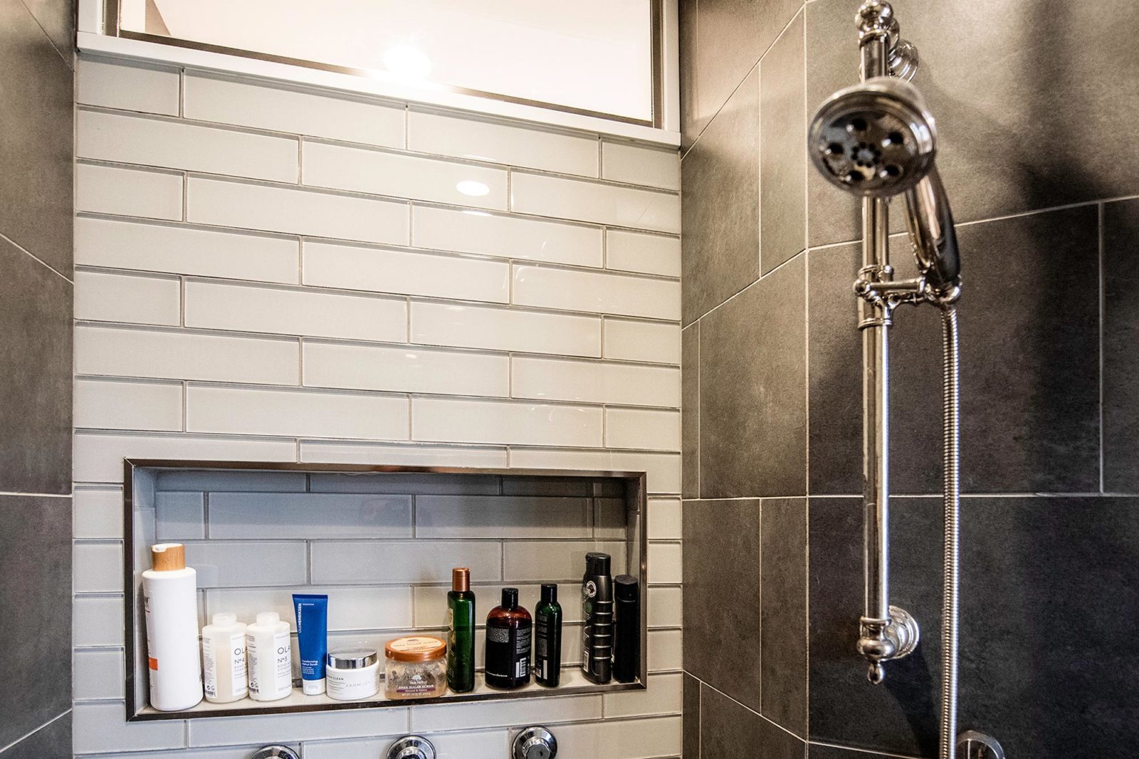 Inside of a shower with white and grey tile walls, silver shower head, and shower products on shelf
