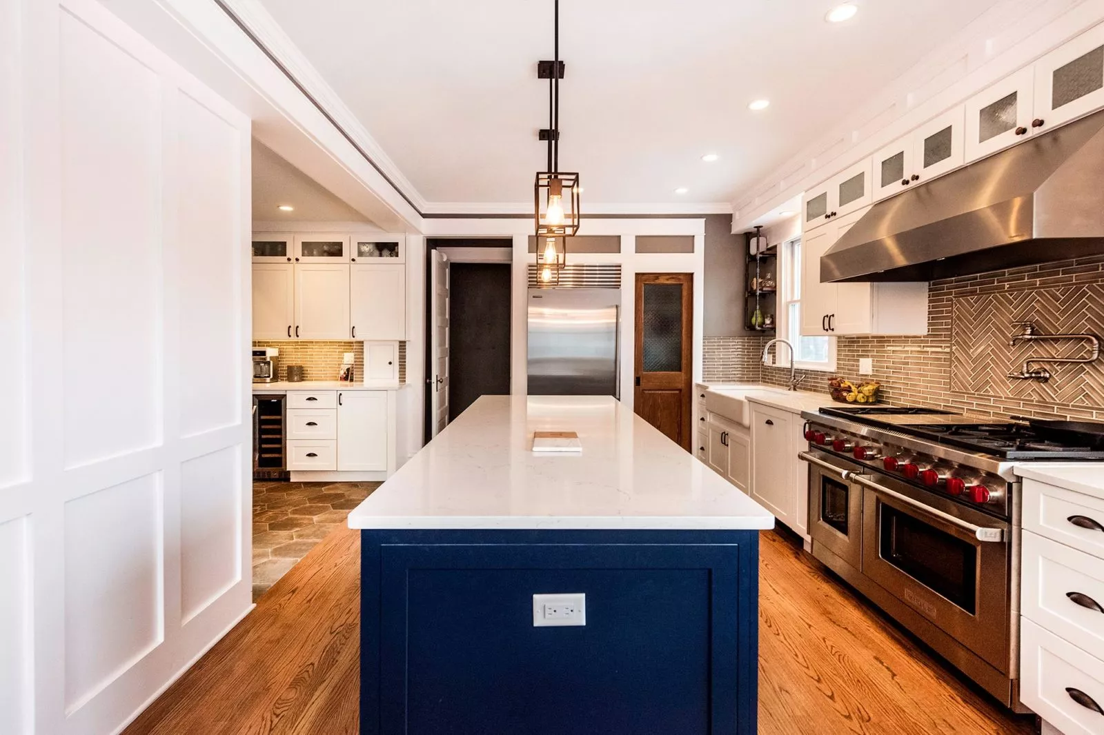 Updated kitchen with black caged iron lights over navy blue white granite-topped island, stainless steel double ovens & fume hood, white cabinets, & brown-tiled statement backsplash