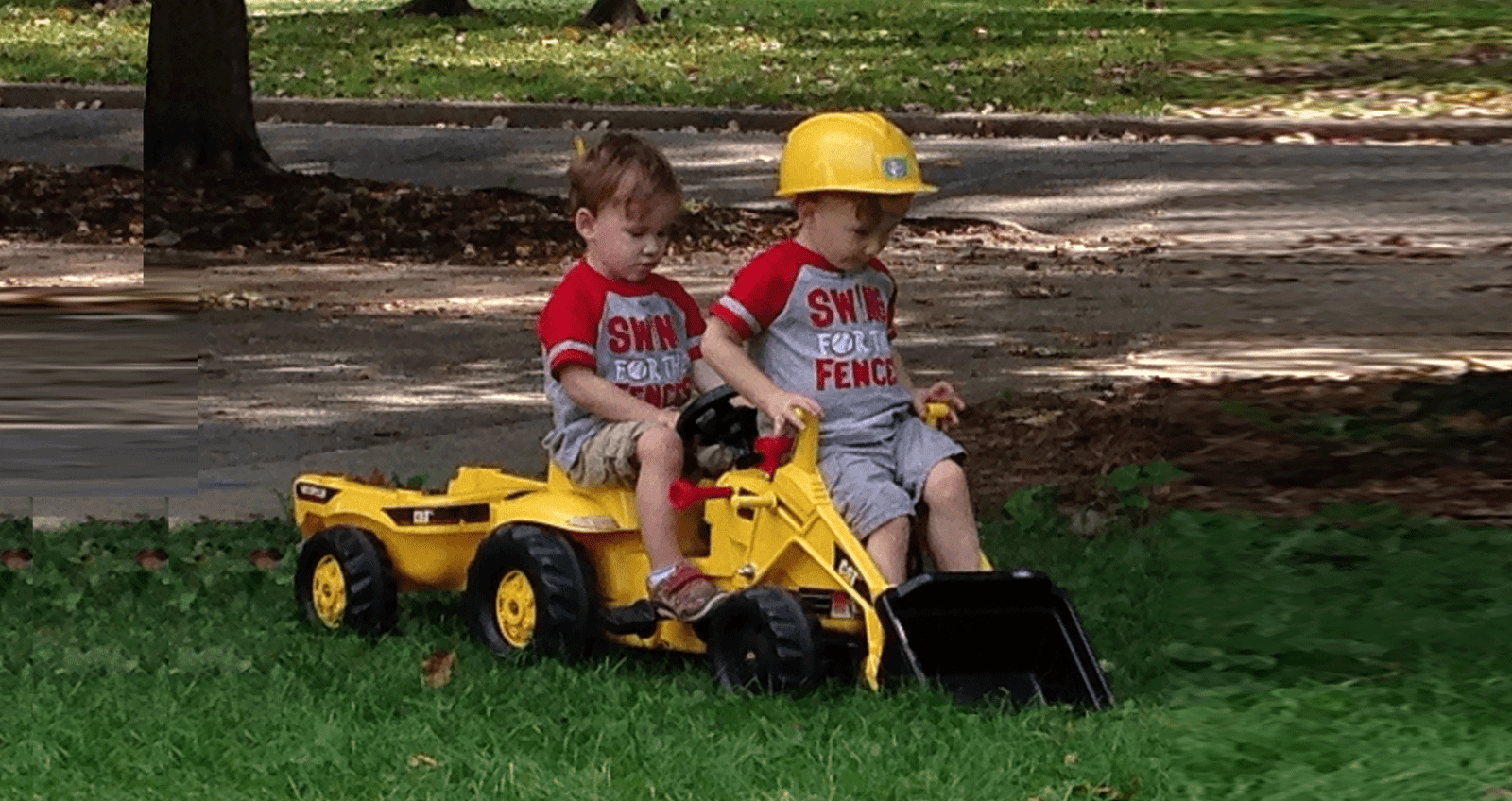 two young children riding on yellow cat tractor toy in green grass next to pavement