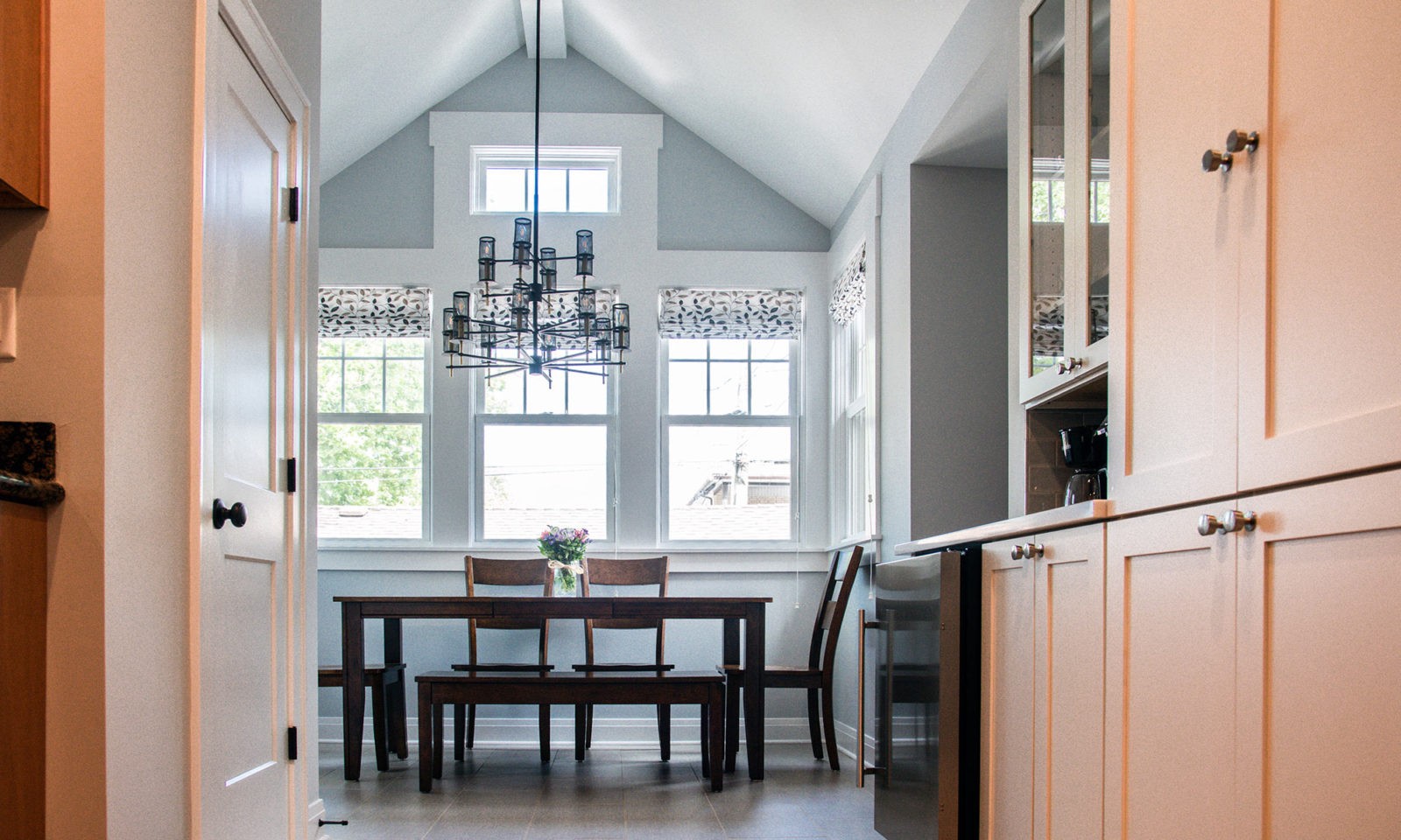 Dark brown kitchen table under a wooden chandelier in an A-frame ceiling kitchen with bright windows on the back wall