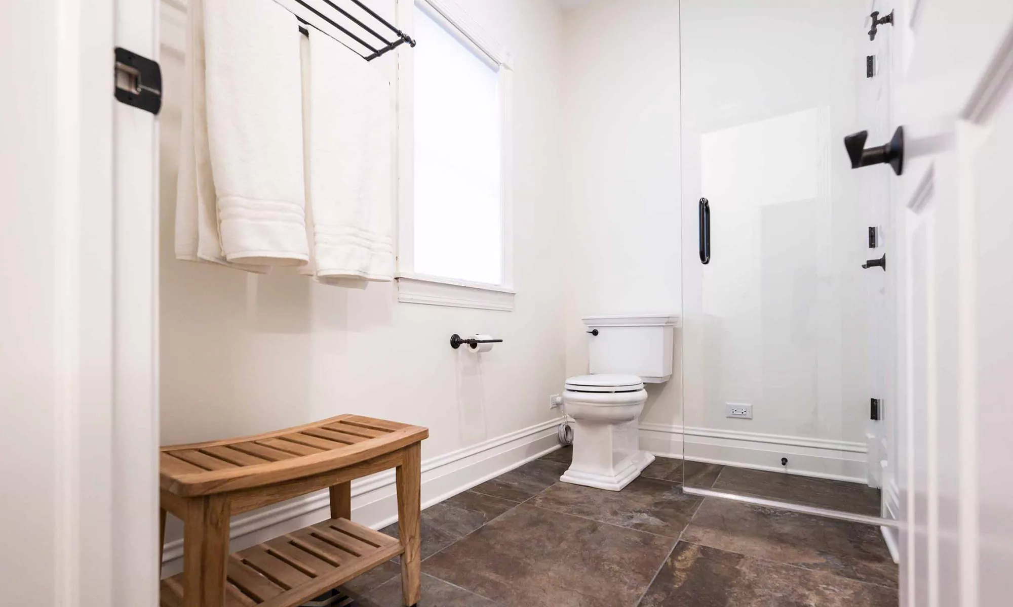 teak bench on grey tile floors in luxury bathroom remodel with toilet in background