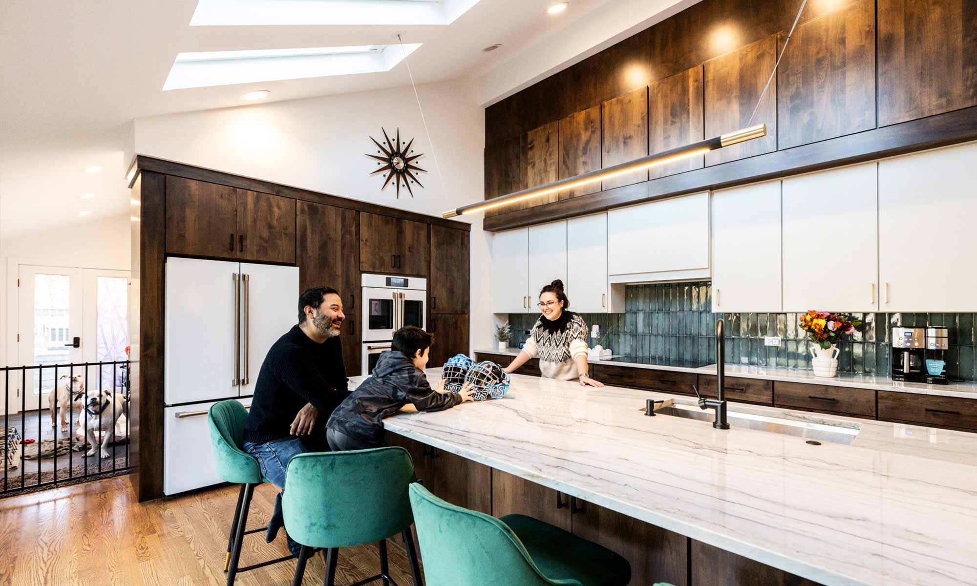 A modern luxury kitchen remodel with dark wood cabinets, white countertops, and a large island with green velvet barstools. A smiling family of three—a man, a woman, and a young boy—gather around the island, engaged in conversation. The boy has hockey gear on the counter, and a sleek pendant light hangs above the island. The kitchen features a skylight, a stylish clock on the wall, and a combination of open and closed cabinetry