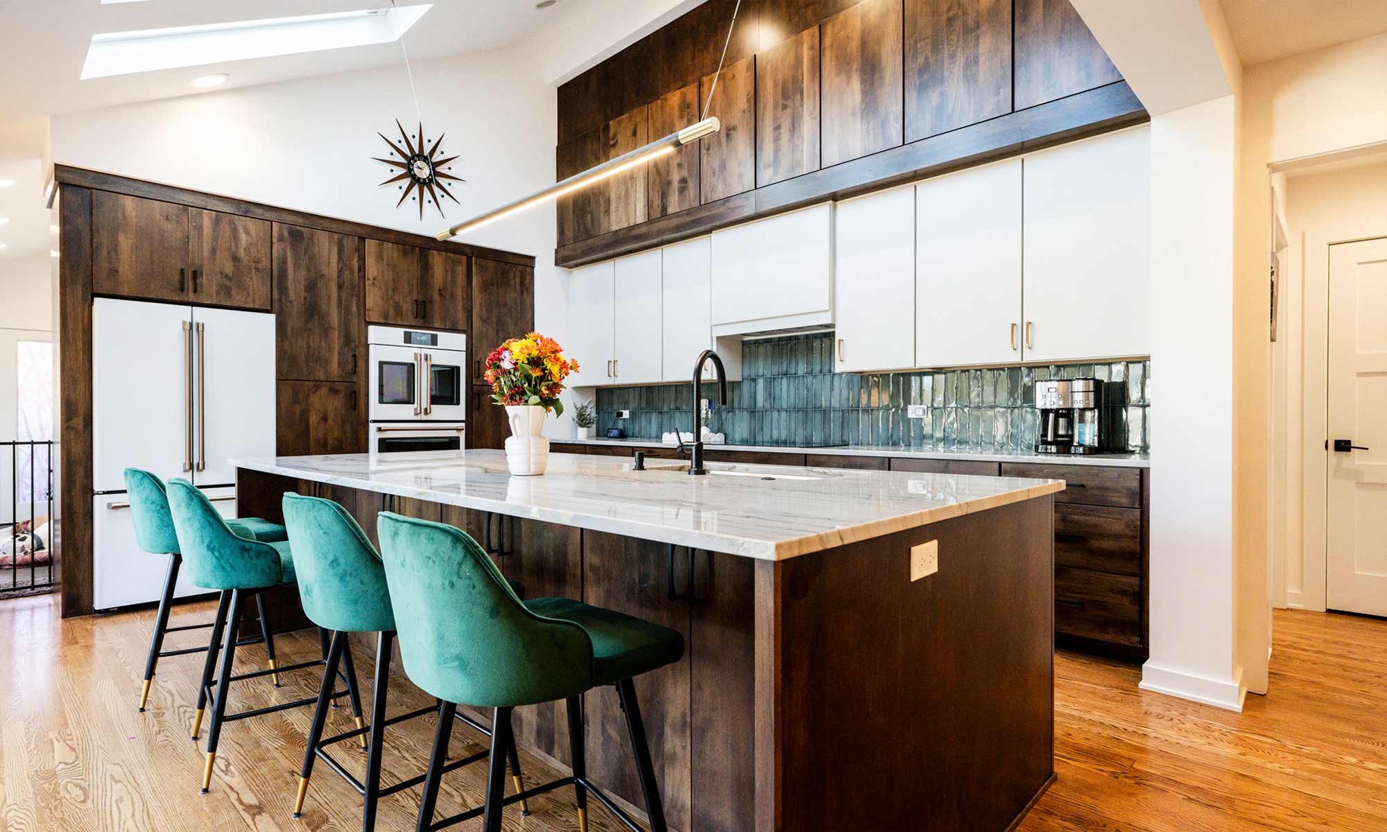 A bright and spacious kitchen with a vaulted ceiling, skylights, and large sliding glass doors. The kitchen has a large marble island with a black faucet and sink. Dark wood cabinets contrast with white upper cabinetry, and a green tile backsplash adds color. Built-in white ovens and a refrigerator are integrated into the cabinetry. Green barstools add a pop of color, while a modern pendant light hangs over the island. A decorative starburst clock is mounted on the wall above the cabinetry.