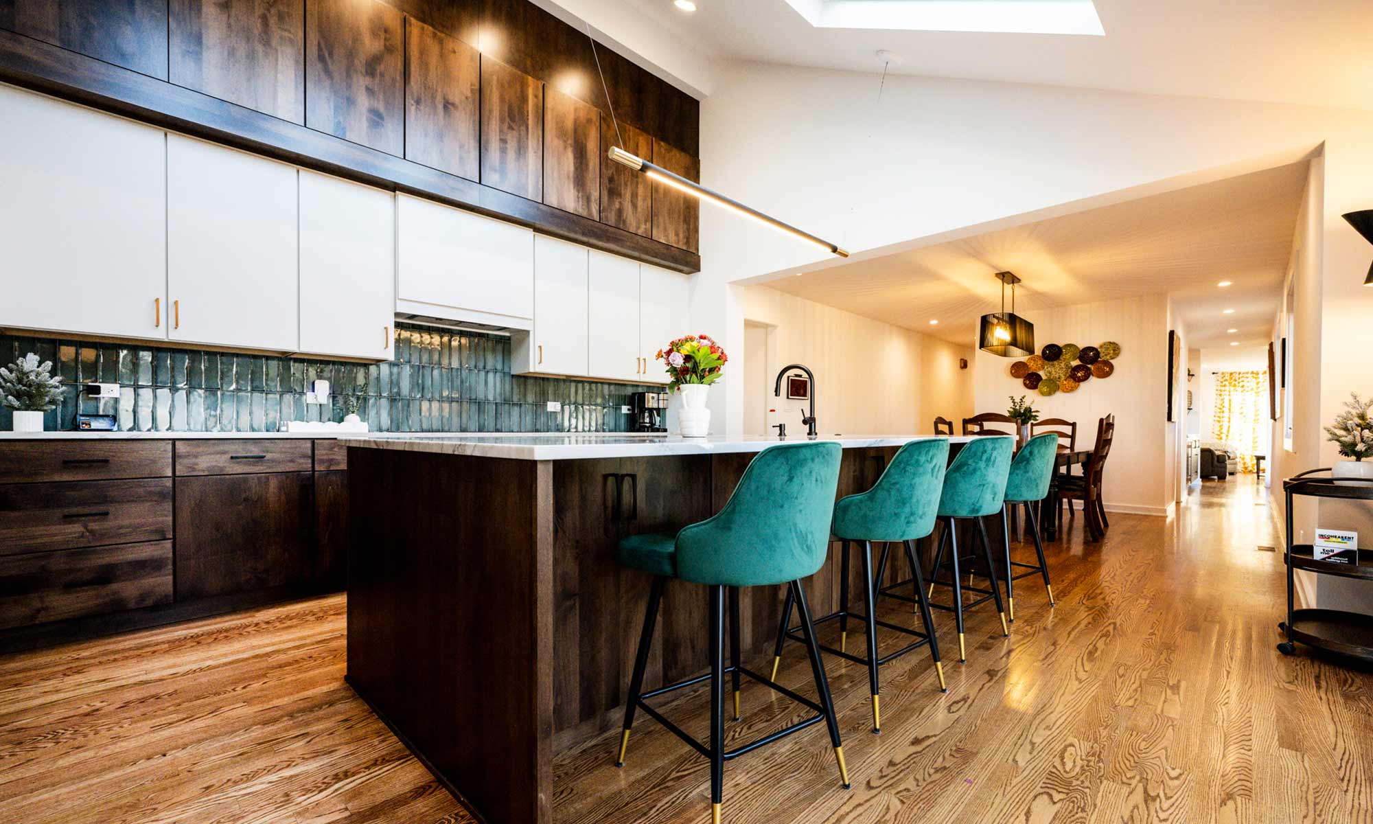 A modern kitchen with dark wood cabinetry and a large marble island. A family of three is gathered around the island, engaging in conversation. Green upholstered barstools line the island, and two dogs are visible behind a gate near the entrance. The space features high ceilings, skylights, and a mix of dark and white cabinets. A black faucet is mounted on the island’s sink, and the backsplash consists of glossy green tiles.