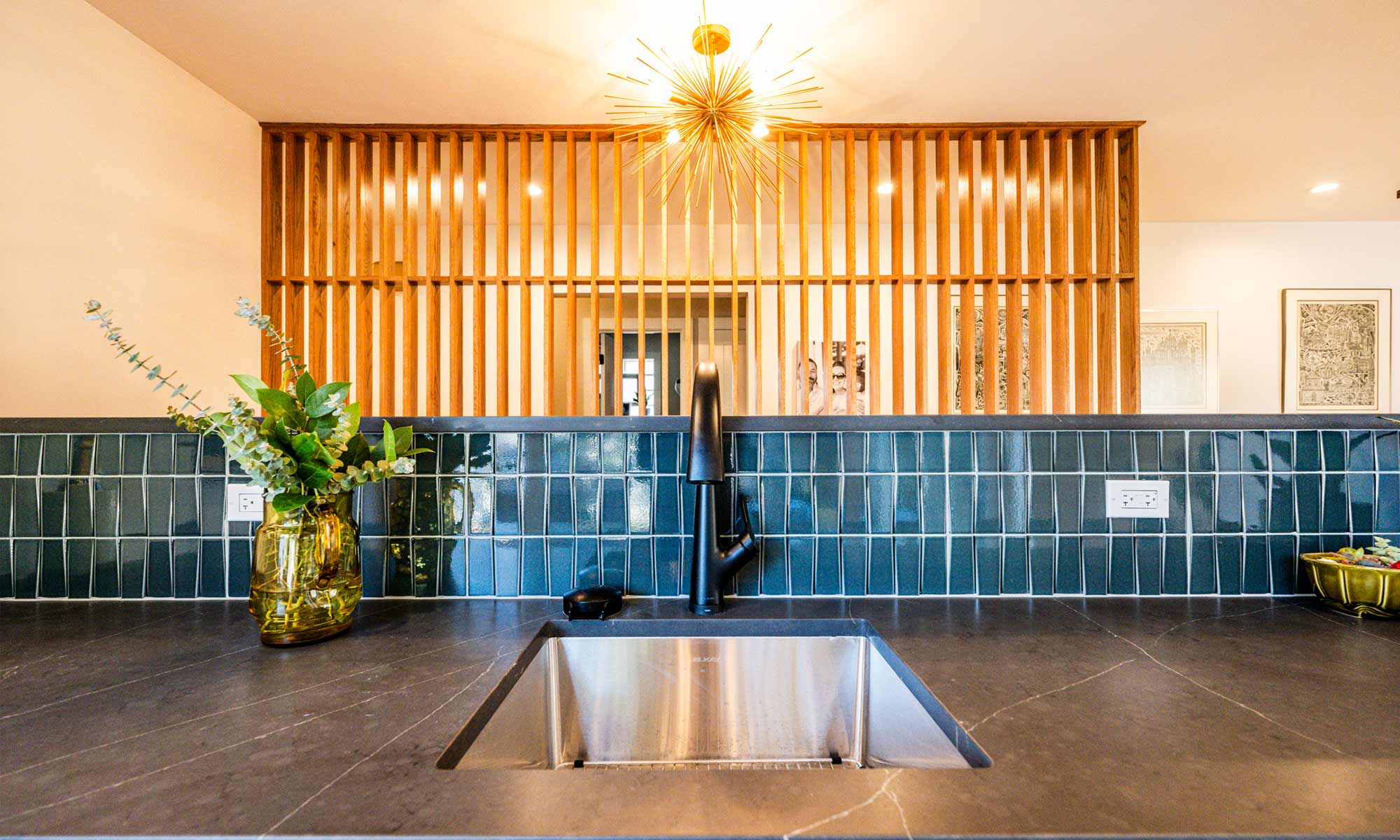 view of a mid-century modern wet bar with wood feature wall beyond, blue backsplash tile and a gold sunburst chandelier