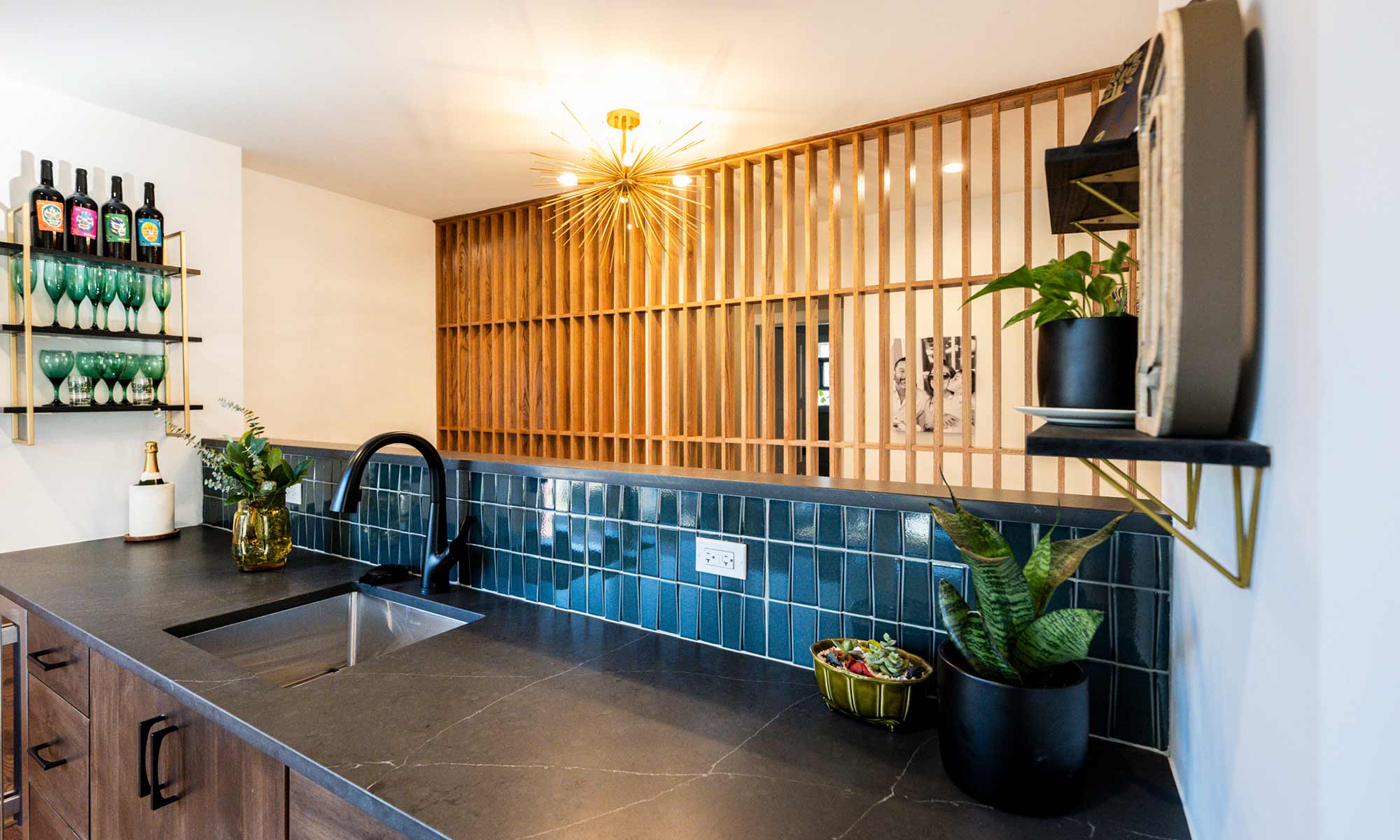 view of a mid-century modern wet bar with wood feature wall beyond, blue backsplash tile and a gold sunburst chandelier