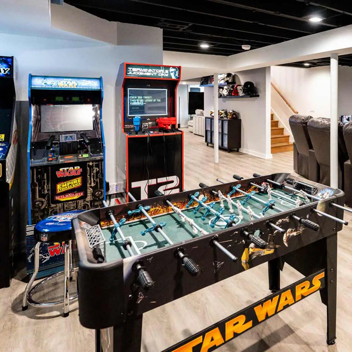 Basement recreation room with Star Wars foosball, vintage arcade cabinets, leather theater seating, and open staircase in the background.