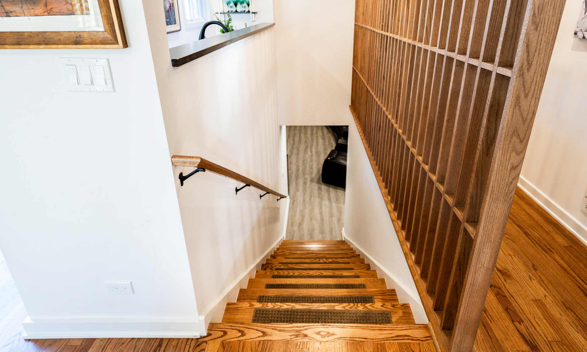 View down a wooden staircase with modern oak railing leading to a finished basement with light-colored vinyl flooring.