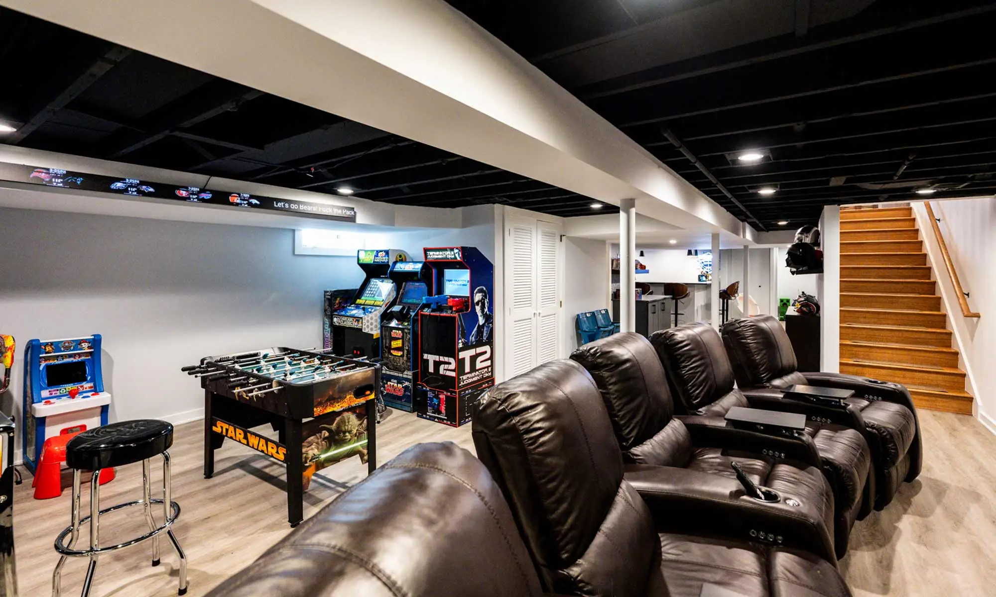 Recreation basement featuring home theater recliners, arcade games, and exposed black-painted ceiling beams.