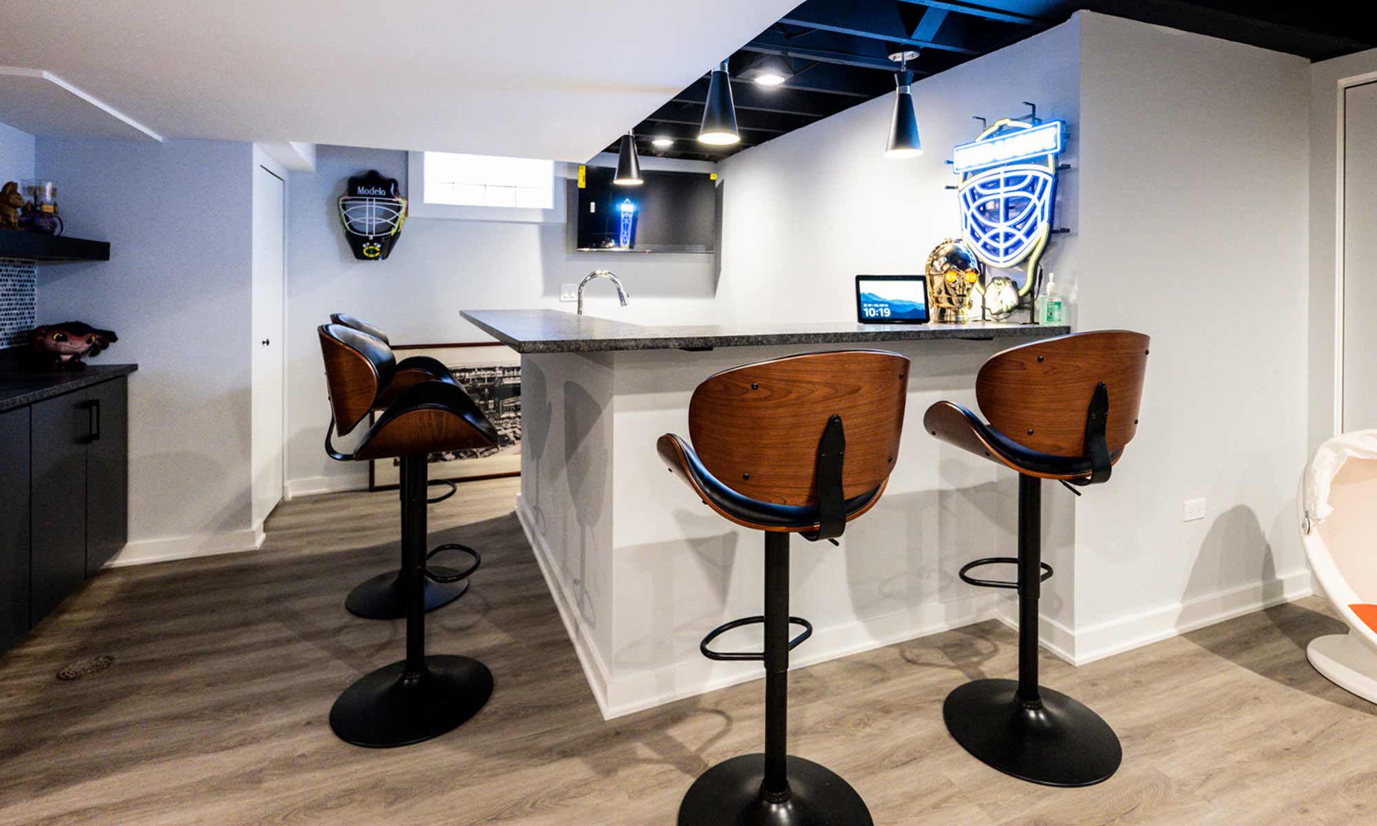 Modern basement bar with three wood and black stools, under-cabinet lighting, and sports-themed neon signs.