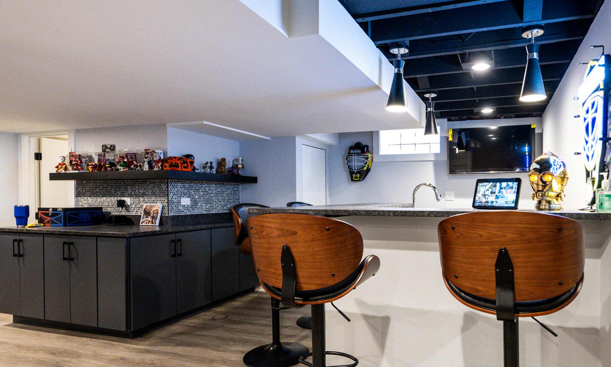 Wide view of a basement bar with toy and memorabilia display shelf, pendant lights, and modern cabinetry.