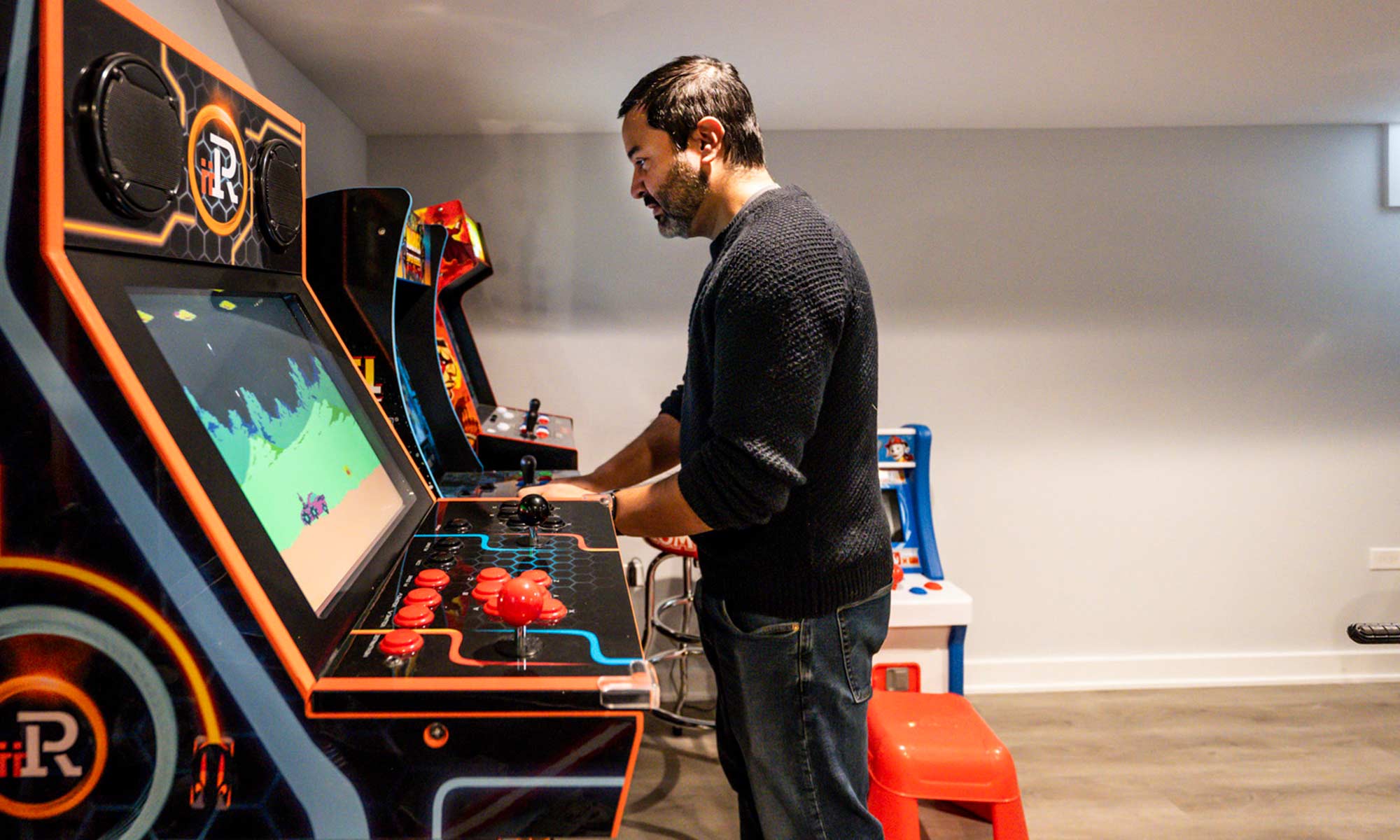 Man playing a retro arcade game in a basement game room lined with vintage machines and kid-friendly game stations.
